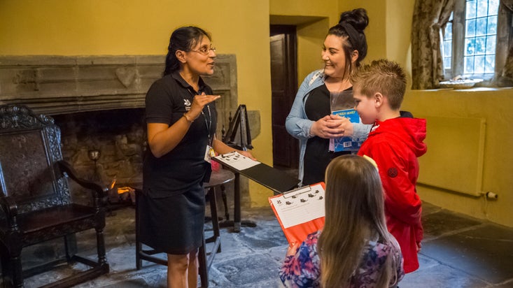 A staff member in a room with a stone fireplace and antique wooden chairs, talks to a family, who are holding clipboards with activity sheets